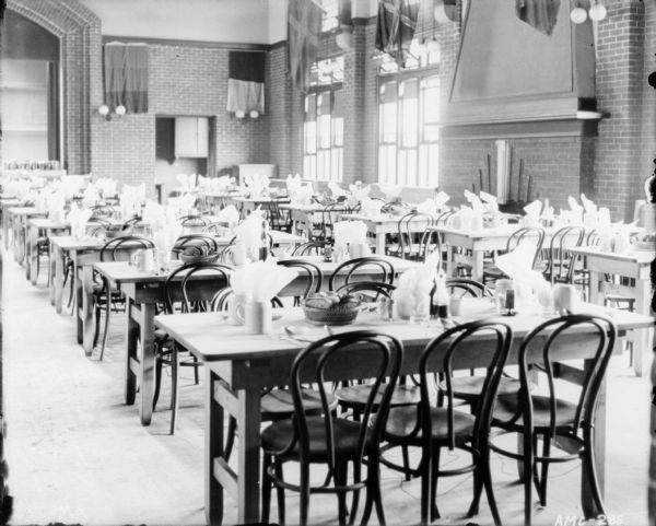 Interior view of dining room. Rows of tables are set with napkins in drinking glasses and mugs, and baskets of bread. Flags hang over the windows on the right and along the back brick wall. The edge of the arched stage at the front of the room is on the left.