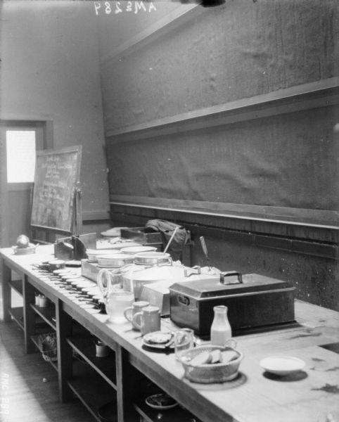 Interior view of the kitchen, with cookware and tableware set out on a long counter. At the back of the room in front of a door is a blackboard.