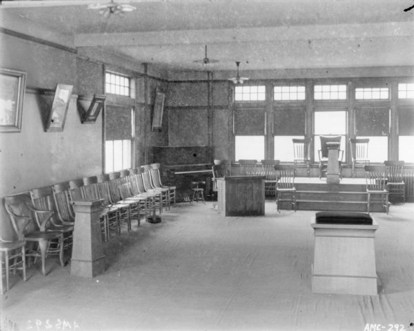 View of lecture hall with chairs along the left wall, a piano in the corner, and more chairs on a raised platform in front of windows at the back of the room.