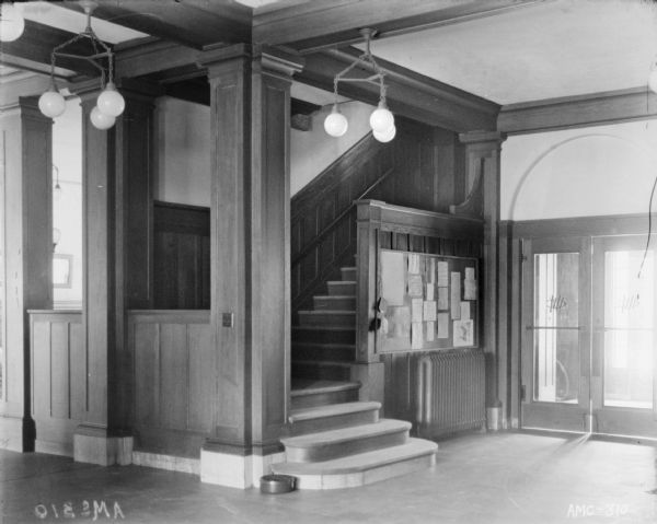Interior view of entry hall of the club house. A spittoon is on the lobby floor at the base of a flight of stairs. A message board is posted near the front door.