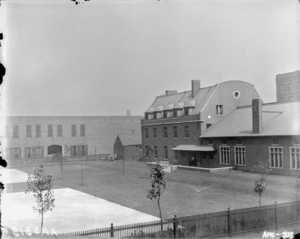 Elevated view over fence of the McCormick Works Club House. There are tennis courts in the left foreground. A man is standing at the door of the club on the right. There is a brick factory building in the background.