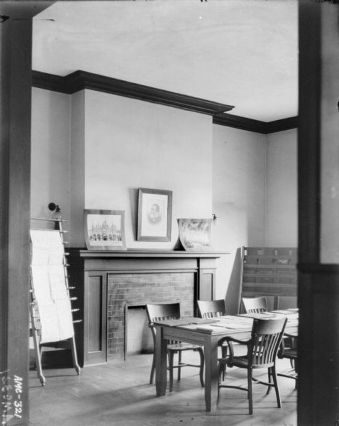 View from doorway of the reading room. In the corner on the left is a newspaper rack. There is a fireplace along the back wall, and on the right is a rack with books. In the center of the room is a table and chairs. A portrait of Cyrus Hall McCormick is above the fireplace.