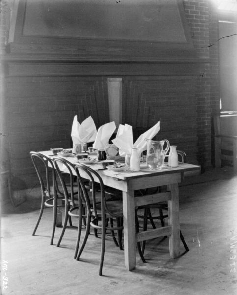 Table in front of large brick fireplace. The table is set with napkins in drinking glasses, mugs, pitchers and baskets of bread.