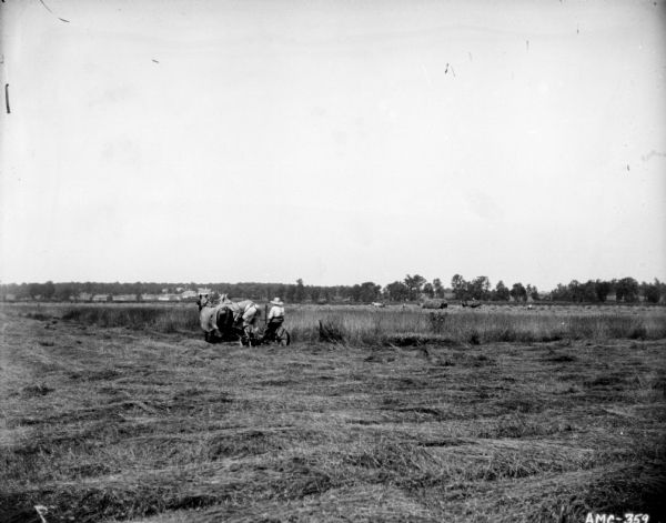 View across mowed field towards man using a horse-drawn mower. In the background on the right people are working near wagons loaded with hay in another field. In the far background are buildings.