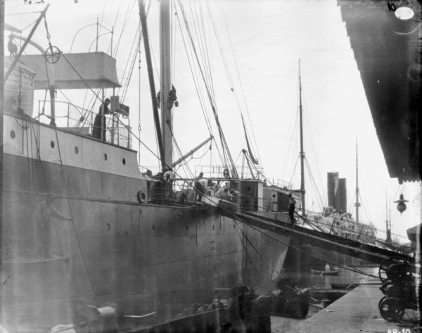 View down dock towards men unloading a binder on the gangway of a ship docked on the left. A woman is standing on the deck of the ship.