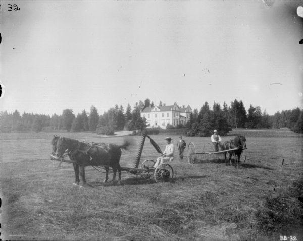 Three men posing in a field at an estate. On the left a man is sitting on a horse-drawn mower. A man in a suit and hat is standing in the center, and on the right a man is sitting on a horse-drawn dump rake. There is a large building in the background.