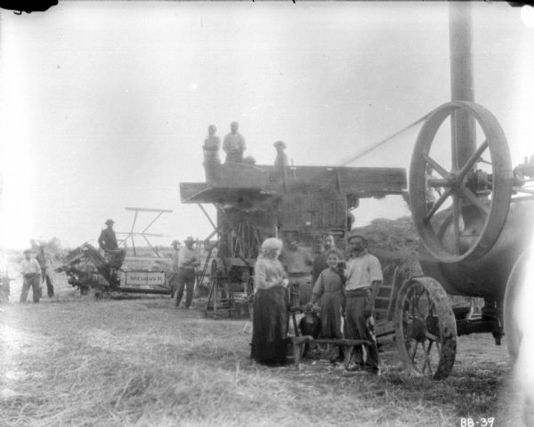 Family posing near farm operation of binders and threshers. In the center a woman is standing with a young girl and a man. Behind them a large group of men are gathered around a McCormick binder and a large threshing machine.