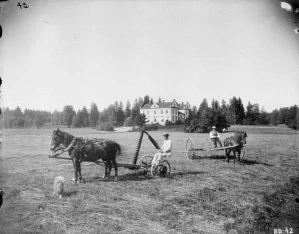 Two men, one driving a horse-drawn mower, and one driving a horse-drawn dump rake, are posing in a large open field with a mansion in the background.