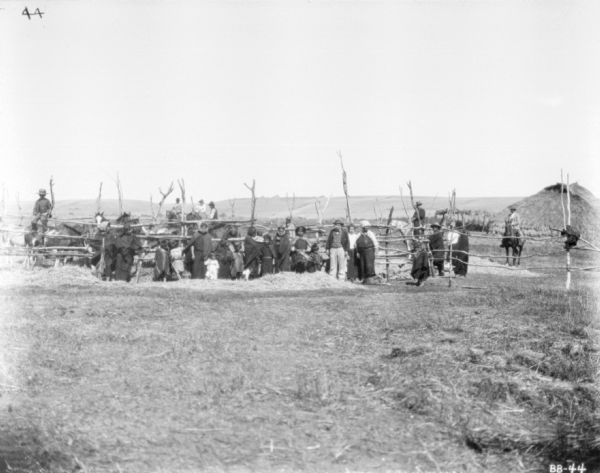 View across field towards a group of men, women and children gathered in front of a corral with horses and oxen. Perhaps Native Americans.