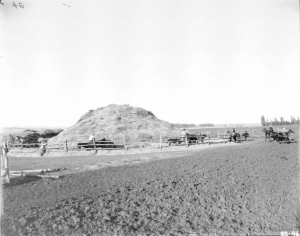 View across field towards a corral with a large haystack inside. Men, some on horseback, are nearby.