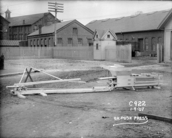 Horse-powered push press and hay press displayed outdoors at Champion Works. Factory buildings are in the background. A small building along the fence has a sign on the side that reads: "No. 1 Hose House."