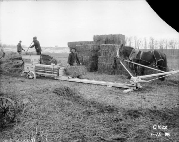 Three men working with a horse-powered hay press. The horse is on the right. There are bales of hay stacked in the background.