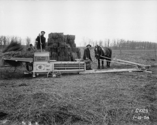 Three men are posing in a field while baling hay with an IHC horse-driven hay press.