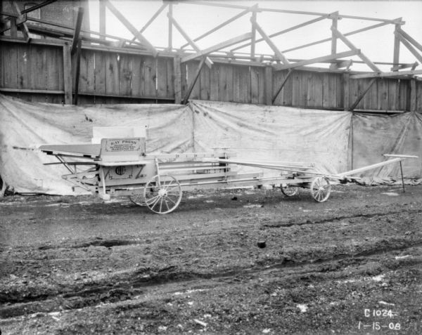 Horse-powered hay press displayed in a yard. There is a white backdrop hanging from a wood wall or fence behind the hay press.