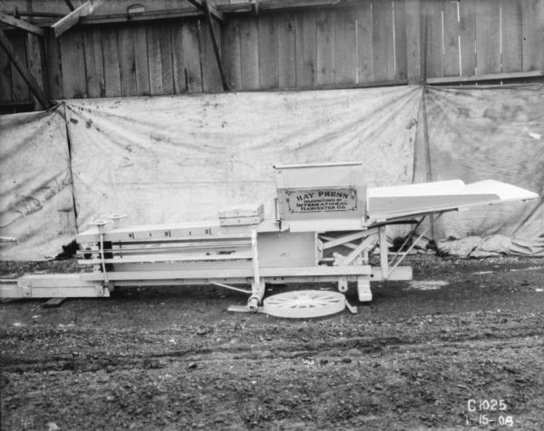 Horse-powered hay press displayed in a yard. There is a white backdrop hanging from a wood wall or fence behind the hay press.