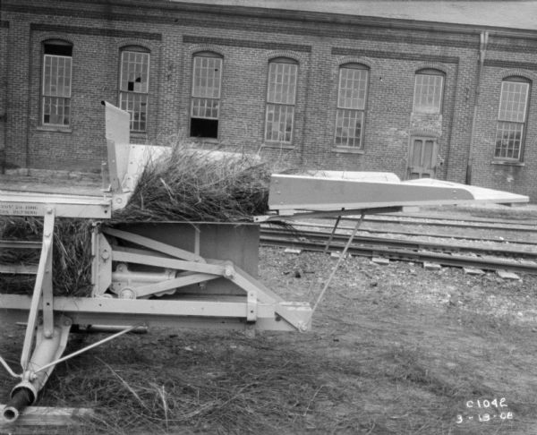 Feeder section of hay press, with hay in the feeder. There are railroad tracks and a brick building in the background.
