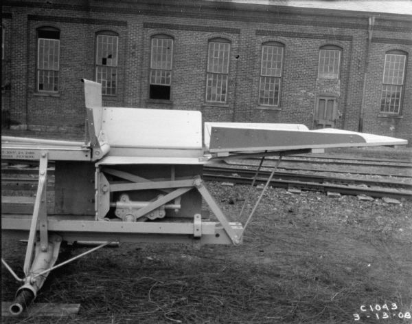 Feeder section of hay press, without hay. In the background are railroad tracks and a brick building.