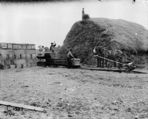 View towards four men using a horse-powered hay press. Three men are working with the hay press, and another man is standing on top of the large haystack. Bales of hay are stacked on the left.
