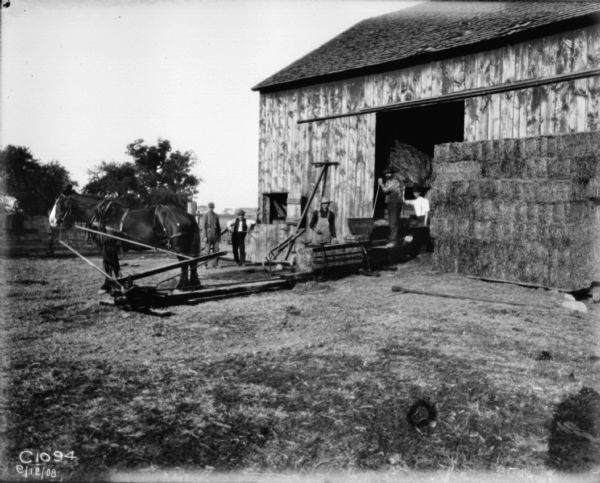 View across barnyard toward men working near an open barn door with a horse-drawn hay press.