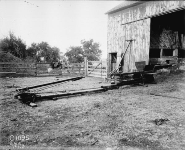 View across barnyard towards a horse-drawn hay press near the open doorway of a barn. In the background is a haystack behind a fence.