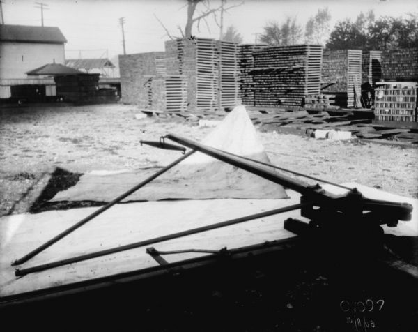 Hay press, in foreground on white cloth, sitting in a yard near stacks of wood or pallets. There are buildings behind a fence in the background.