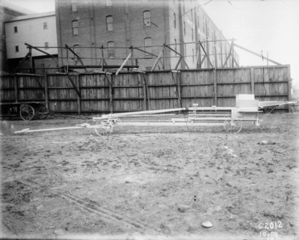 Side view of hay press in yard. In the background is a wagon near a fence, and large brick buildings beyond. On the side of the brick building is a painted sign that reads: "International."