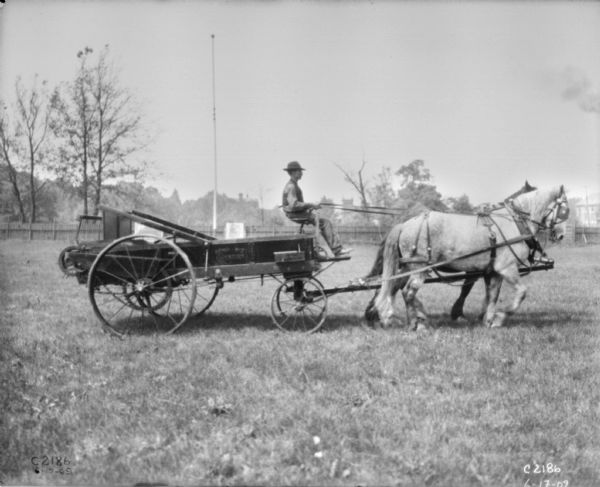 Right side view of a man driving a horse-drawn manure spreader. In the background is a fence.