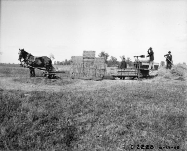 View across field towards three men using a horse-drawn hay press to bale hay. One man is standing on a pile of hay on the right. Another man is standing on the hay press, and another man is standing behind the hay press near stacked bales of hay.