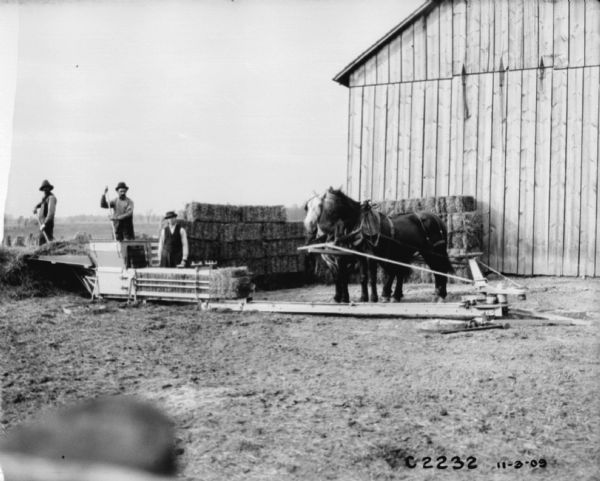 View towards three men operating a horse-powered hay press near a barn.