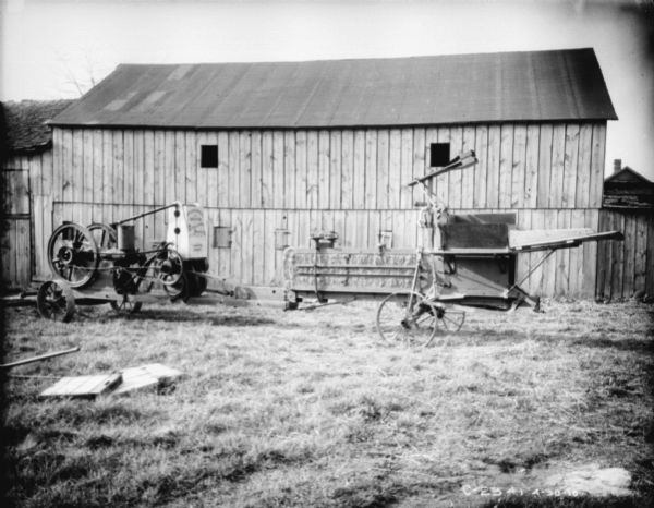View across yard of powered hay press in front of a barn at Springfield Works, with bales of hay in the machine.