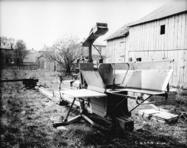 Three-quarter view from loading end of powered hay press. There is a barn on the right, and in the background behind a fence are what may be houses.