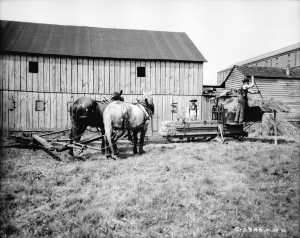 Two men operating horse-powered hay press in front of a barn. There is a factory building in the background on the right.
