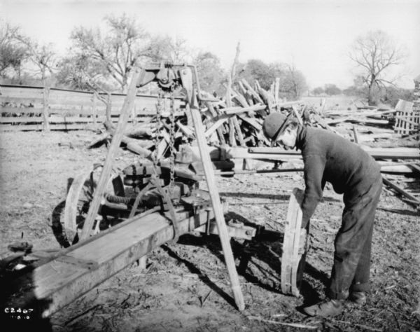 Man outdoors operating lifting jack for a hay press. There is a large pile of wood in the background and a fence.