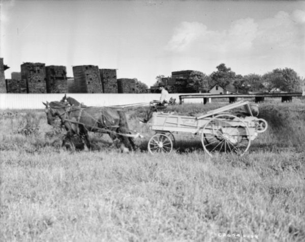 View across field towards a man using a horse-drawn manure spreader. In the background is a fence and beyond are tall stacks of wood or pallets, and buildings among trees.