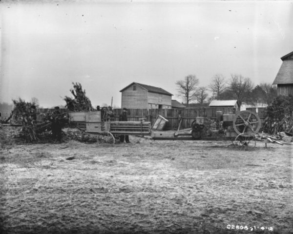 View towards men using a hay press to bale cornstalks. There is a fence and farm buildings in the background.