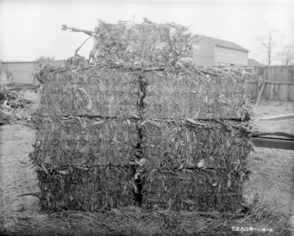 Stock of corn bales. In the background are buildings behind a fence.