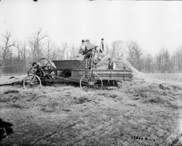 Men in a field using a powered hay press. The belt-driven engine is out of the frame to the left. One man is standing on top of a haystack, and three men are working near the hay press.