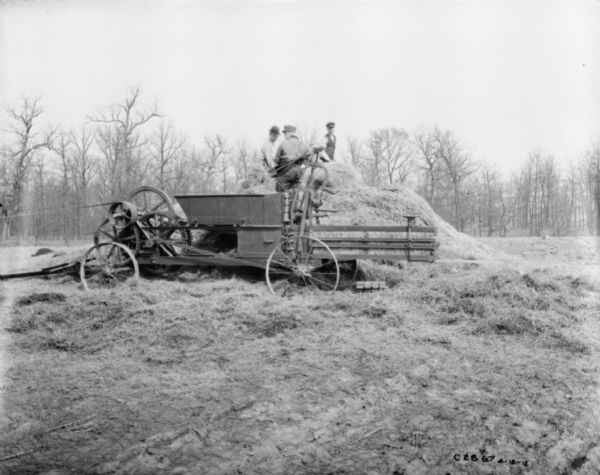 Men in a field using a powered hay press. The belt-driven engine is out of the frame to the left. One man is standing on top of a haystack, and two men are working near the hay press.