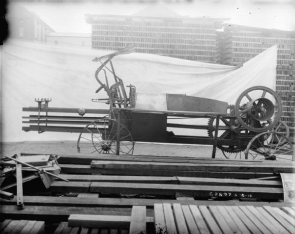Hay press set up in front of a white sheet used as a backdrop. Behind the sheet are stacks of wood and factory buildings.