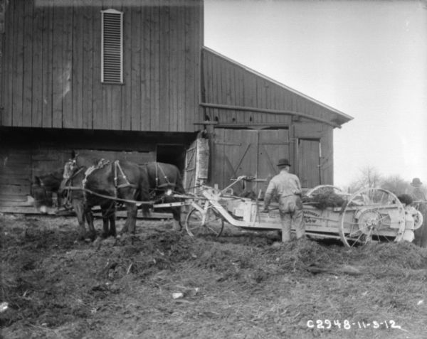View towards men loading a horse-drawn manure spreader near a barn.