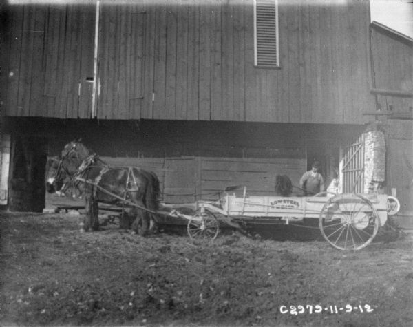 View towards a man loading a horse-drawn manure spreader near a barn.