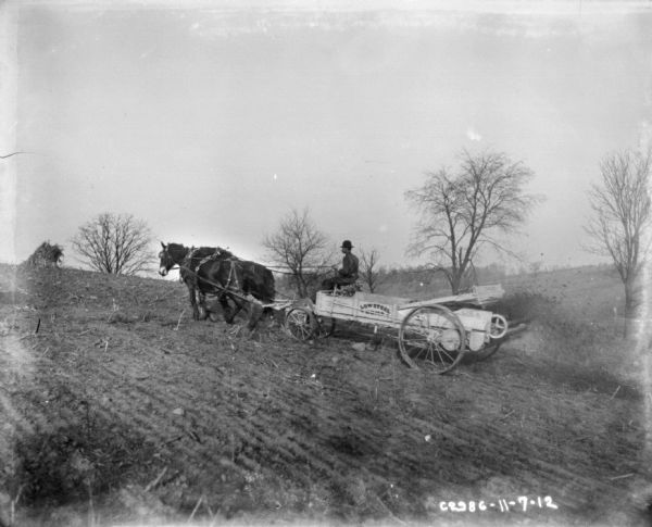 Left side view of a man using a horse-drawn manure spreader in a field on a hill.
