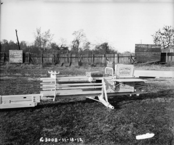 Hay press in yard. The side of the press has "IHC Press Made By International Harvester Co.," and "Nov. 20, 1906, Aug. 31, 1909" painted on the side. In the background is a fence, which has a billboard advertising "Everbody's Print Shop."
