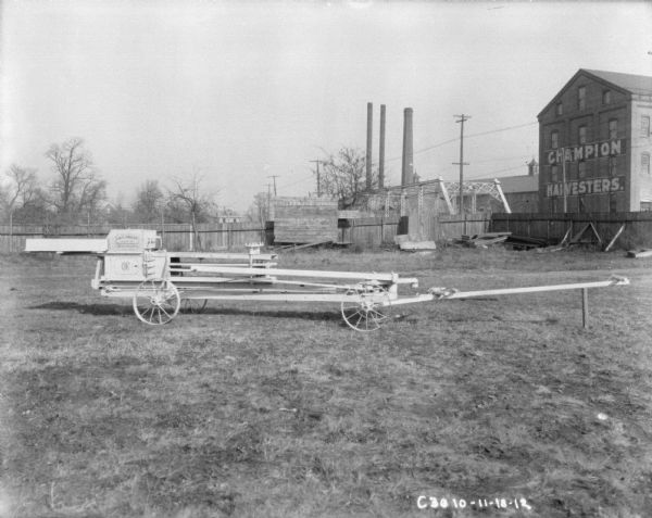 Hay press in yard. In the background is a fence. The side of the press has "IHC Press Made By International Harvester Co.," painted on the side. In the background are smokestacks and factory buildings behind a fence, and one is painted with a sign for "Champion Harvesters."