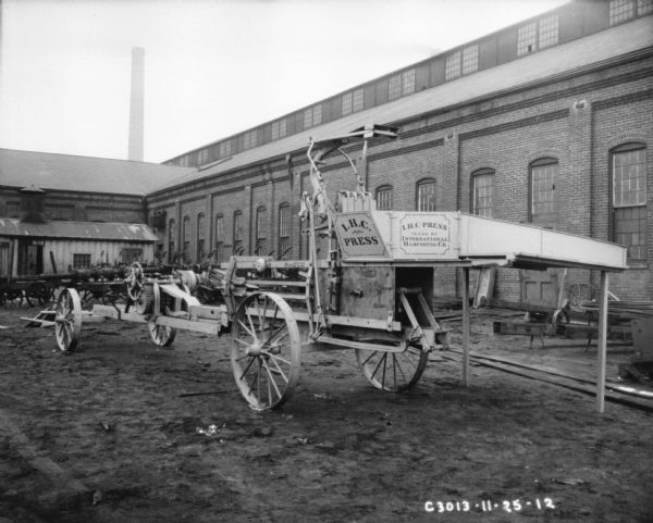 Hay press outdoors at Springfield Works. The side of the press has "IHC Press Made By International Harvester Co.," painted on the side. Factory buildings are in the background.