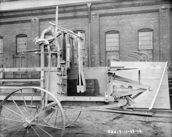 Powered hay press outdoors. There is a large brick factory building in the background.