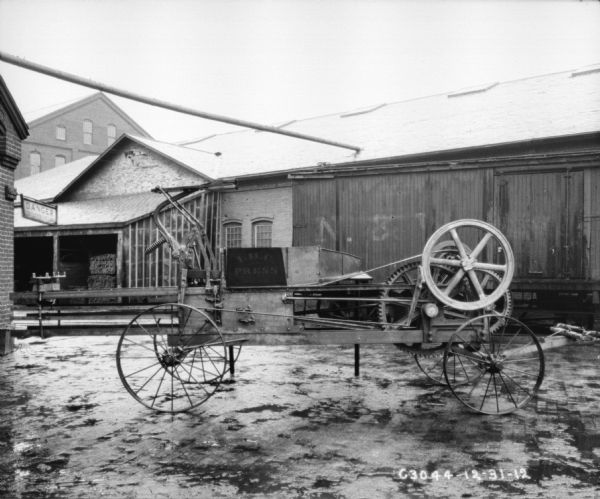 Powered hay press outdoors in yard. In the background is a railroad car and factory buildings.