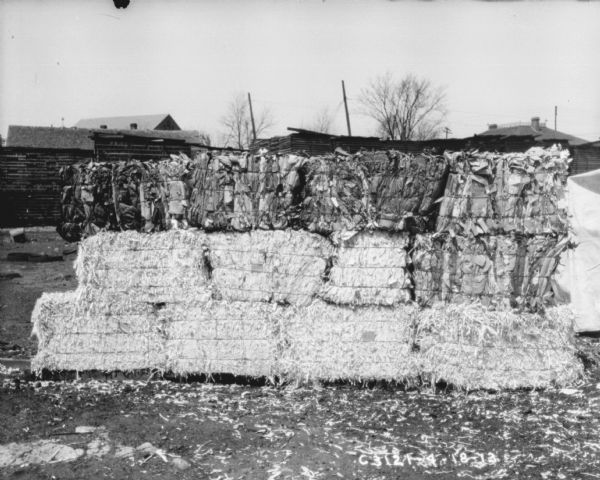 Stacks of hay bales in a yard. In the background are stacks of lumber or pallets, and buildings.