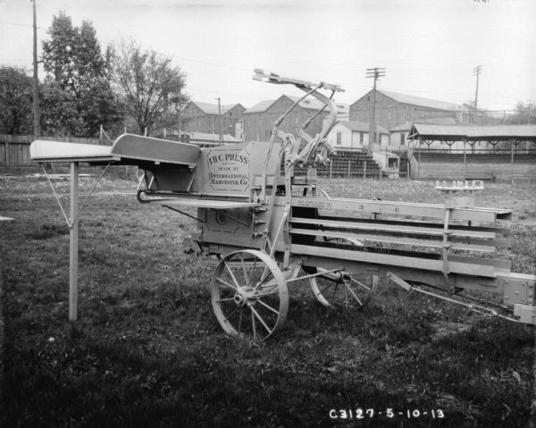 Hay press outdoors in fenced yard. There is a fence and factory buildings in the background. The side of the press has "I.H.C. Press" painted on the side.