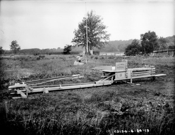 Hay presses outdoors sitting in long grass at Springfield Works. The hay press in the foreground has "I.H.C. Press" painted on the side. In the background are billboards against a fence. There is a break in the fence, and on the right the fence is sagging.